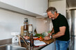 © VALENTINA BARRETO STUDIO/Westend61 - Smiling mature man cutting vegetable while standing in kitchen at home