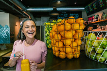 Smiling Mature Woman Holding Fresh Juice In Mason Jar While Standing By Oranges In Metallic Basket At Bar