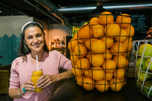 Smiling Mature Woman Holding Fresh Juice In Mason Jar While Standing By Oranges In Metallic Basket At Cafe