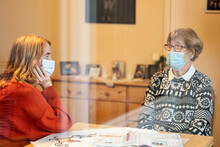 Granddaughter And Mother Wearing Face Mask Talking While Sitting With Social Distance At Home During Covid-19