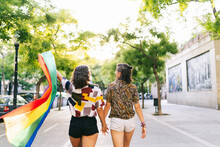 Lesbian Couple With Rainbow Flag Holding Hands While Walking On Footpath