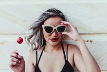 Beautiful Woman Wearing Sunglasses While Holding Red Lollipop Against Wall