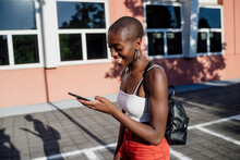 Cheerful Young Woman With Shaved Head Using Smart Phone While Walking On Sidewalk In City
