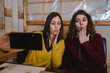 © Pintau Studio - Two beautiful young women taking a selfie photo posing giving a kiss and making grimace faces in a restaurant or cafe. Friendship, togetherness concept.