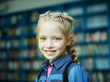 © Comeback Images - Closeup head and shoulders portrait of cute blonde little girl with braided hair and freckles smiling happily at camera standing near book shelves in school library