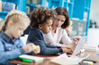© Comeback Images - Side view selective focus of female teacher explaining task on laptop screen to mixed race elementary student, his classmates studying sitting at desk in school classroom nearby