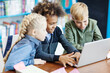 © Comeback Images - Side view of three curious diverse students with laptop sitting at desk in elementary school classroom. Mixed  race schoolboy playing computer game, his friends supporting him