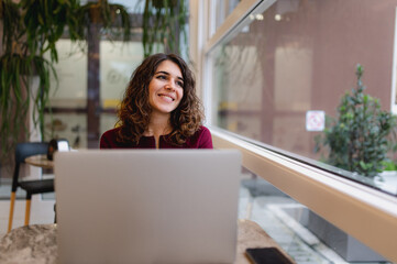 Close-up portrait of young woman in a cafe sitting at a table using computer laptop. Lady smiling while looking out the window.