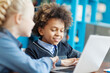 © Comeback Images - Side view of two smiling kids, boy and girl, using laptop computer together sitting at desk in school library. Focus on curly-haired mixed race male student