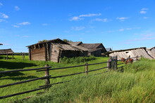 The Old Wooden Village Buildings Were Dilapidated And Collapsed Behind A Fence Of Long Cross-sticks On Bright Green Grass On A Sunny Summer Day With Blue Skies. Picturesque Landscape Of Rural Life