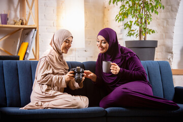 Wall Mural - Tea time. Happy and young two muslim women at home talking, smiling, having fun, sitting on sofa. Friendship, modern tech, emotions concept. Lifestyle and culture, arabian traditions, ethnicity.