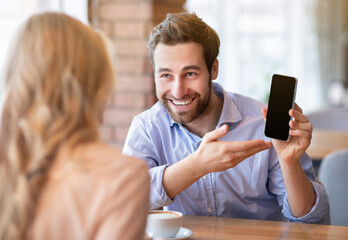 Positive millennial guy showing his girlfriend cellphone with blank screen at coffee shop, mockup for design