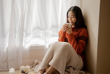 Young Beautiful Asian Woman Holding Cup Of Coffee And Marshmallows On Top, Sitting At Home And Looking Out The Window. Happy Girl Drinking Chocolate In Sweater In Cold Weather Winter