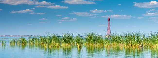 Wall Mural - panoramic view to the old red metal lighthouse on the river in summer day under blue sky with clouds and copy space