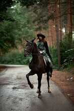 Young Beautiful Woman In A Suit, Green Dress, Corset, Black Topm Hat On A Black Horse On Nature In The Forest. Fairy Tale, Creative Photo Session Of A Girl With A Horse