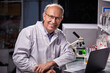 © Roman - researcher in medical suit posing at vamera in laboratory, successful scientist work on laptop, with test tubes on table, smiling
