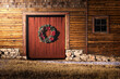 A Christmas wreath hangs on a wood barn door. There is a little snow on the ground. There is also a window off to the right and some stonework at the bottom of the barn. There is wood texture.