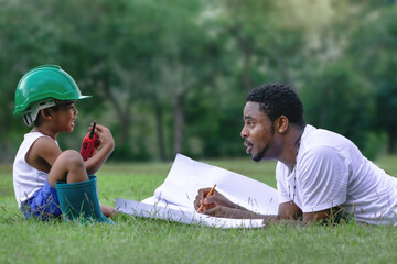 Side view of African father explained the construction with blue print to his son, lying on lawn at park, son in safety hat and walkie talkie or portable radio in hand