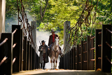 Two young cowboys and a horse cross a wooden bridge.