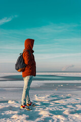 Back view of male tourist with rucksack standing on coast of the frozen bay while journey. Man traveler wearing red jacket with backpack explore nature. Wanderlust lifestyle.
