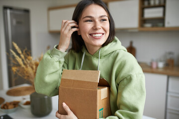 Shopping, delivery and purchase concept. Indoor image of joyful fashionable young woman holding cardboard box, unpacking furniture she ordered for new apartment, having excited happy facial expression