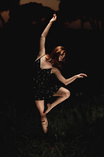 Elegant Female Ballet Dancer In Dress Standing In Meadow With Raised Arms And Performing In Dark Park In Iceland