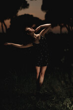 Elegant Female Ballet Dancer In Dress Standing In Meadow With Raised Arms And Performing In Dark Park In Iceland