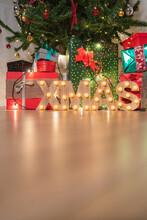 Christmas Tree With Wrapped Presents And Wooden Letters Of Word Xmas With Light Bulbs In Cozy Room