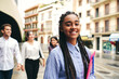 © Fatima Guisado/ADDICTIVE STOCK - Young smiling woman with Afro braids and plastic folder looking at camera behind multiracial coworkers