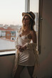 © Gustavo Pozo/ADDICTIVE STOCK - Young seductive female in stylish apparel and straw hat standing looking away on windows in apartment