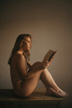 Side View Of Sensual Female Wearing Lace Underwear Sitting On Wooden Table With Book And Enjoying Story