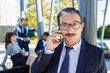 © Ignacio Ferrandiz/ADDICTIVE STOCK - Positive adult male manager in formal outfit and eyeglasses talking on smartphone while resting on bench in park with unrecognizable diverse colleagues during break