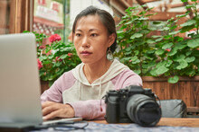 Concentrated Asian Female Journalist Sitting At Table With Digital Photo Camera And Using Laptop While Preparing New Article