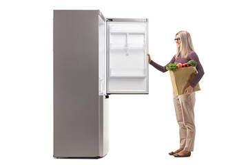 Wall Mural - Young woman holding a bag of groceries in front of an open fridge