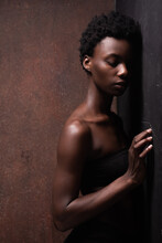 Seductive African American Female Model Wearing Black Top With Bare Shoulders Leaning Against The Wall And Closed Eyes During Photo Session In Dark Studio