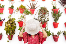 Anonymous Female Tourist Covering Face With Straw Hat Near White Wall With Different Potted Plants