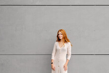 Positive Young Female With Long Red Hair Wearing Casual Dress And Smart Bracelet Looking Away While Standing Against Concrete Wall