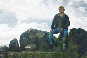 A resting bearded hiker in yellow hat relaxing on a rock and looking far away. Metallic steel thermos, backpack and plants on the foreground. Beautiful clear sky and clouds in the background.