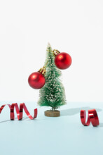 Miniature Green Christmas Tree Decorated With Red Baubles Placed On Table With Ribbons In Studio On Blue Background