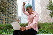 © David Munoz/ADDICTIVE STOCK - Side view low angle of excited African American male entrepreneur rejoicing at success in project while sitting with laptop and raised fist on street