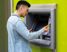 Side View Of Young Asian Male With Smartphone In Hand Inserting Card Into ATM Terminal For Withdrawing Cash On City Street