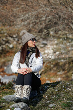 Female Tourist In Warm Boots And Coat Sitting On Ground In Woods On Sunny Day While Embracing Knees And Relaxing During Trekking In Winter