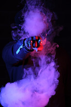 Anonymous Stylish Male Showing Fist With Rings In Shape Of Skull And Covering Face In Cloud Of Smoke Illuminated By Purple Neon Light In Dark Studio