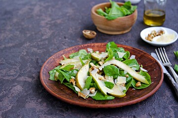 Green salad of fresh spinach, juicy pear, walnuts and parmesan on a brown clay plate on a dark concrete background.