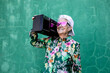 © Sergio Victor Vega/ADDICTIVE STOCK - Cool elderly female in colorful blouse and trendy cap and sunglasses carrying record player and enjoying music against green wall