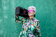 © Sergio Victor Vega/ADDICTIVE STOCK - Cool elderly female in colorful trendy blouse and sunglasses carrying record player and enjoying music against green wall