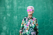 © Sergio Victor Vega/ADDICTIVE STOCK - Confident senior gray haired female in stylish colorful clothes and trendy sunglasses smiling happily while standing against green wall