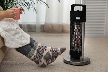 Canvas Print - Woman with tea warming legs near halogen heater at home, closeup