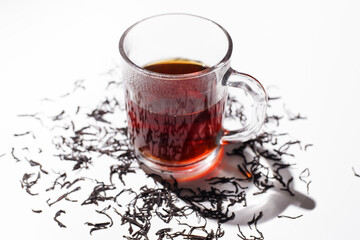 Glass cup of black tea and scattered dry ceylon tea leaves on white table, close up