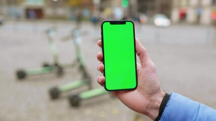 Busy young man holding phone with green screen smartphone touching and swiping outdoors on the street background electric scooters. Internet message mock-up display. Slow motion